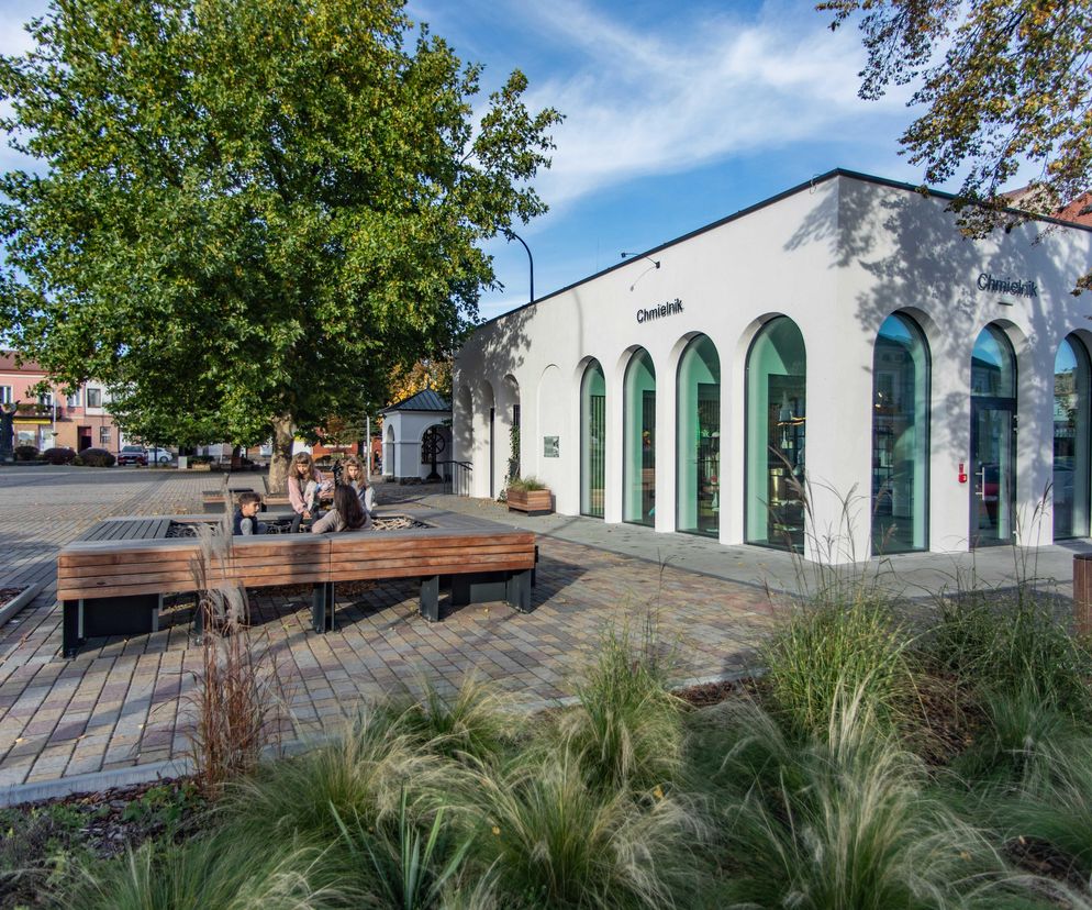 Zrewitalizowany Rynek w Chmielniku, zaprojektowany przez Gdak Studio, z nowoczesnym pawilonem z łukowymi przeszkleniami. Na pierwszym planie drewniane ławki i ozdobna roślinność, a także grupa osób. W tle fragment ulicy oraz kamienice. Więcej o projekcie na portalu Architektura Murator Plus.