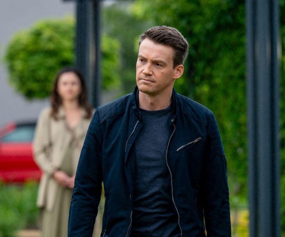 Aktor Krzysztof Kwiatkowski jako Jakub w ciemnej kurtce, patrzący z powagą w prawo, z Pauliną Lasotą jako Kasią w tle. Kontekst zdradza, że Jakub wybaczy Kasi zdradę w 1880 odcinku M jak miłość. Więcej o tym na Super Seriale.