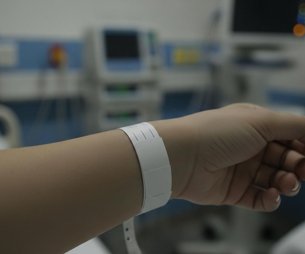 Ludzka ręka, prawdopodobnie należąca do pacjenta, znajduje się w centrum kadru, rozciągając się od lewej dolnej do prawej środkowej części zdjęcia. Na nadgarstku widać białą opaskę identyfikacyjną z pionowymi liniami i dwoma widocznymi perforacjami. Dłoń jest lekko zaciśnięta, z widocznymi zagięciami skóry na kostkach. Tło jest rozmyte, ale rozpoznawalne jako wnętrze szpitala, z widocznymi fragmentami niebieskich i białych ścian, a także niewyraźnymi urządzeniami medycznymi z ekranami i lampkami.
