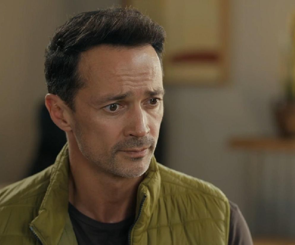 Aktor Rafał Cieszyński jako Mariusz z serialu Barwy szczęścia w zielonej kamizelce, z zamyślonym wyrazem twarzy, nawiązujący do problemów z byłą narzeczoną, o czym przeczytasz więcej na portalu Super Seriale.