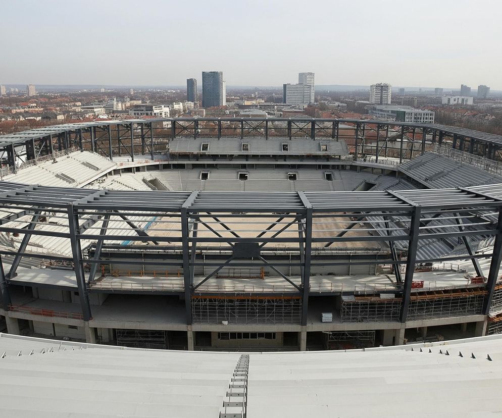 Panoramiczny widok ukazuje duży, owalny stadion w trakcie budowy, z częściowo wykończonymi trybunami w odcieniach szarości i rozległą, szarą konstrukcją wsporczą dachu, składającą się z masywnych stalowych belek. Wokół stadionu rozciąga się miejski krajobraz z licznymi budynkami mieszkalnymi o czerwonych dachach i kilkoma wyższymi wieżowcami w tle. Horyzont dominuje jasne, nieco zamglone niebo.