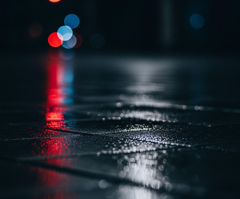 Czarna, mokra powierzchnia, przypominająca chodnik lub drogę, rozciąga się od dołu obrazu, zwężając się ku górze. Na jej powierzchni widoczne są liczne, drobne kropelki wody, błyszczące w świetle. W tle, rozmyte czerwone i niebieskie światła tworzą efekt bokeh, odbijając się jednocześnie w wilgotnej nawierzchni.