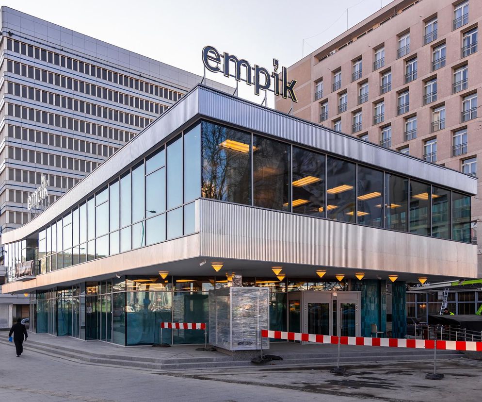 Budynek handlowo-usługowy z zaokrąglonymi narożnikami i dużymi przeszklonymi witrynami na parterze, widoczny na tle wyższych biurowców. Nad wejściem widnieje neon Empik. Przed budynkiem, gdzie pawilon Cepelii jest w remoncie, stoją czerwono-białe barierki, a po prawej stronie widać podnośnik. O architekturze Warszawy przeczytasz na Architektura Murator Plus.