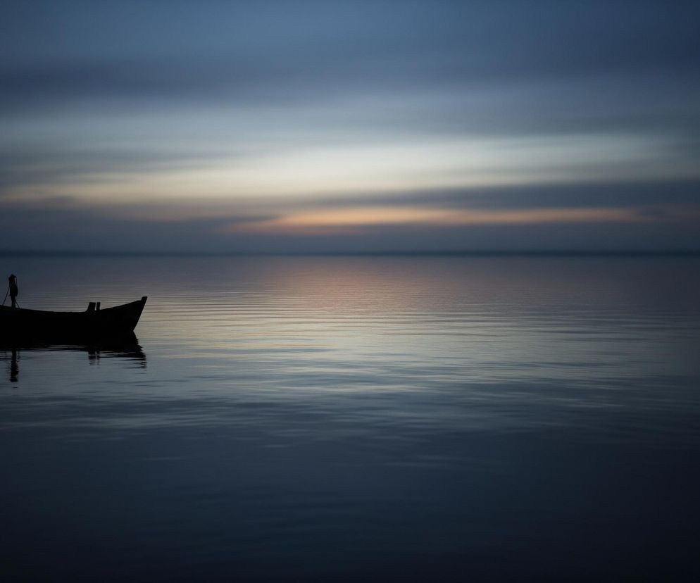 Czarna, niewielka łódka z dziobem skierowanym w lewo, unosi się na spokojnej tafli wody w dolnej części kadru. Jej ciemna sylwetka odbija się w wodzie, tworząc wyraźny refleks. Horyzont oddziela gładką, ciemnogranatową wodę od rozmytego nieba, które przechodzi od ciemnego granatu u góry, przez odcienie szarości i błękitu, aż do jasnego, brzoskwiniowo-różowego paska tuż nad linią wody. Całość utrzymana jest w chłodnych, stonowanych barwach.