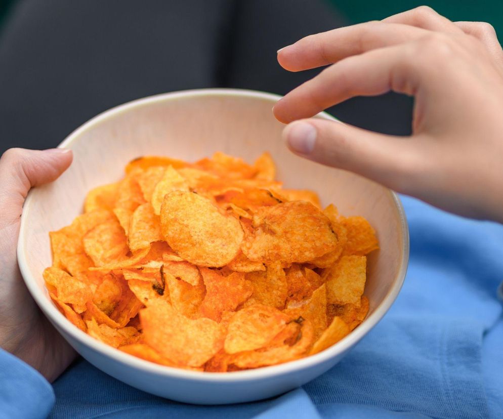 Osoba w niebieskiej bluzie trzyma miseczkę z pomarańczowymi chipsami, a druga dłoń sięga po przekąskę. Kontekst dotyczy szkodliwości przetworzonej żywności i tego, jak przestać jeść chipsy. Więcej na ten temat przeczytasz na portalu Poradnik Zdrowie.