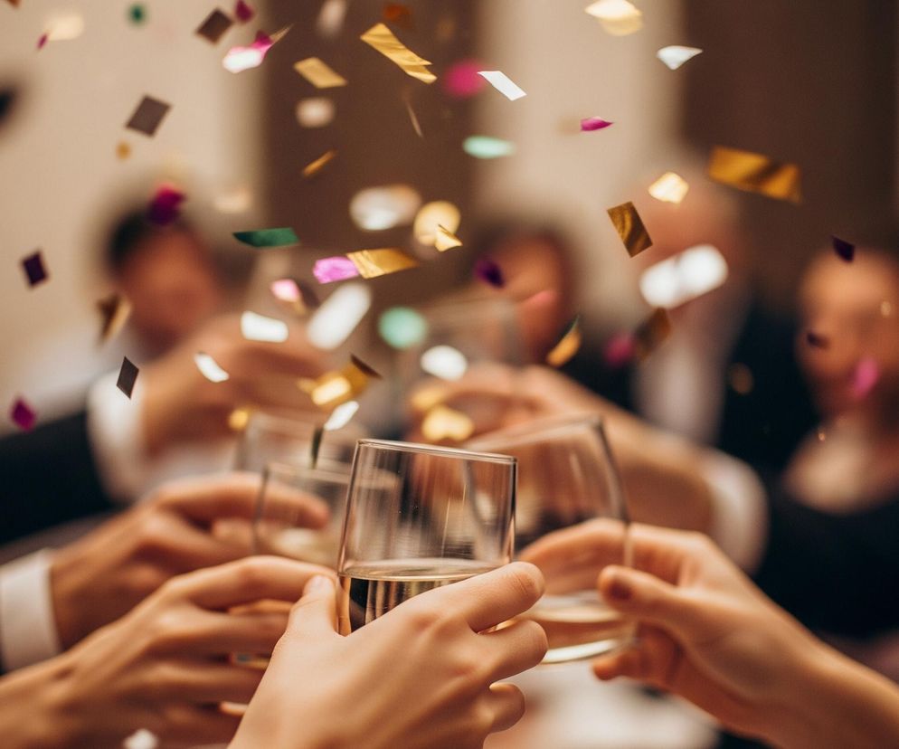 Grupa ludzi wznosi toasty szklankami z jasnym napojem, prawdopodobnie szampanem lub białym winem, przy czym dłonie z kieliszkami stanowią centralny, wyostrzony element obrazu. W tle widoczne są rozmyte sylwetki innych osób, niektóre ubrane w ciemne garnitury z białymi kołnierzykami. Nad sceną unosi się deszcz kolorowego konfetti w odcieniach złota, fioletu, różu, bieli i zieleni, co dodaje zdjęciu świątecznego charakteru. Całość utrzymana jest w ciepłej tonacji brązowo-złotawej.