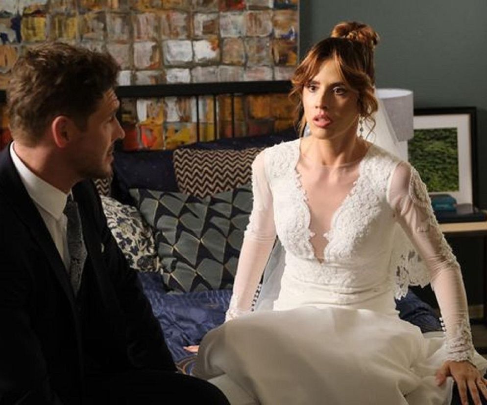 Michalina Sosna jako Kama w białej sukni ślubnej, z wyraźnym zdziwieniem na twarzy, rozmawia z Mikołajem Roznerskim w roli Marcina. Scena z serialu M jak miłość ilustruje opóźnienie ślubu przez sen Kamy. Więcej informacji o serialu znajdziesz na Super Seriale.