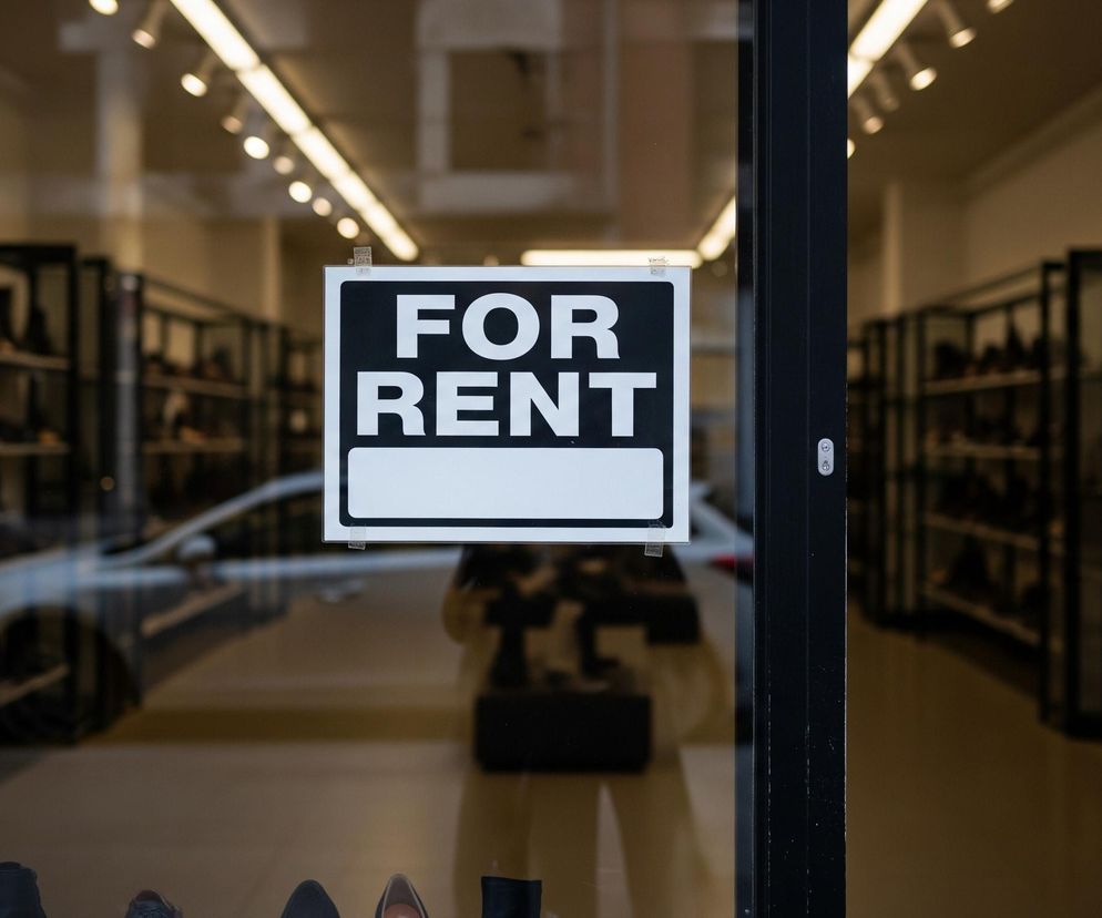Na pierwszym planie, centralnie, wisi biała tabliczka z czarnym napisem FOR RENT (DO WYNAJĘCIA) na górze, poniżej znajduje się pusty biały prostokąt. Tabliczka jest przyklejona do przeszklonych drzwi lub okna. W odbiciu na szybie widać fragment ulicy, w tym samochód w srebrnym kolorze, a także niewyraźną sylwetkę osoby fotografującej. Przez szybę widać wnętrze sklepu obuwniczego z wieloma półkami, na których stoją różne buty. W tle widoczne jest oświetlenie wnętrza, składające się z wielu podłużnych i punktowych lamp.