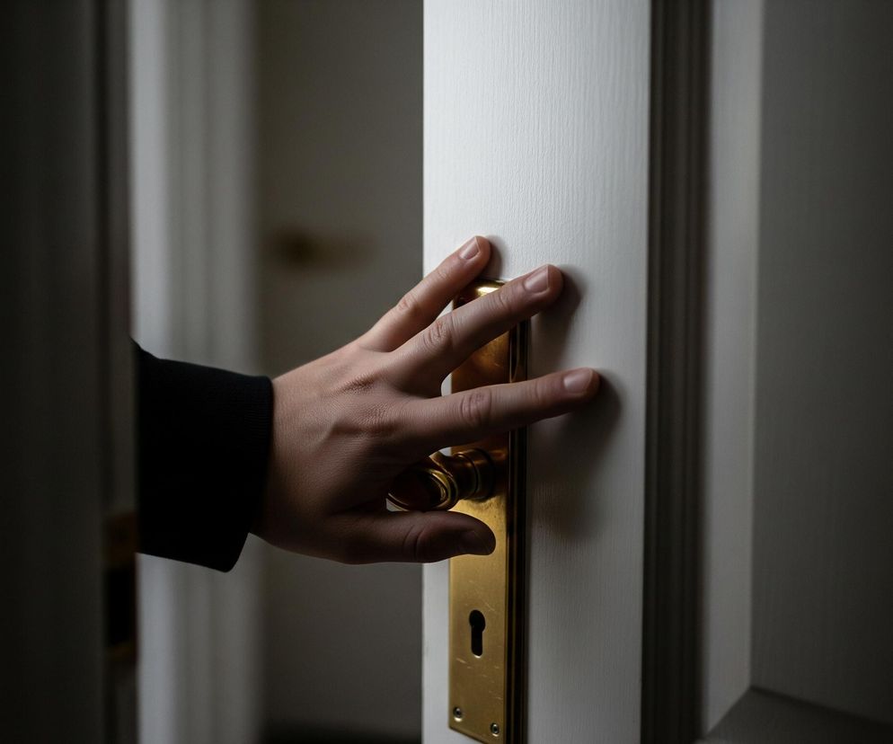 Na zdjęciu widoczna jest ludzka dłoń, ubrana w ciemny rękaw, dotykająca złotej klamki na białych drzwiach. Klamka ma owalny uchwyt i prostokątną płytę z dziurką od klucza. Scena jest słabo oświetlona, z lewej strony kadru dominuje cień, a po prawej widać krawędź otwartych drzwi, za którymi rozciąga się ciemne pomieszczenie.