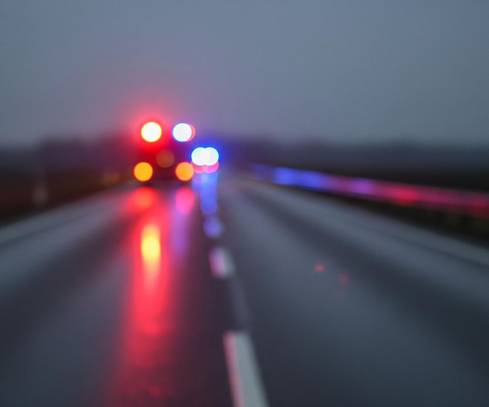 Przedstawiony obraz to rozmyte ujęcie drogi w nocy lub o zmierzchu, z intensywnymi, jaskrawymi światłami w oddali. Na pierwszym planie widoczna jest mokra, ciemna nawierzchnia jezdni, z wyraźnymi białymi liniami rozdzielającymi pasy ruchu, które odbijają światło. W centrum kadru, w oddali, znajdują się okrągłe, rozmyte plamy światła: kilka intensywnie czerwonych i pomarańczowych po lewej stronie oraz jasnoniebieskie i białe po prawej, sugerujące światła pojazdów uprzywilejowanych. Po prawej stronie drogi widoczna jest rozciągająca się bariera ochronna, której powierzchnia również odbija czerwone i niebieskie światło. Całe tło jest ciemnoszare, lekko zamglone, z niewyraźnymi konturami linii horyzontu.