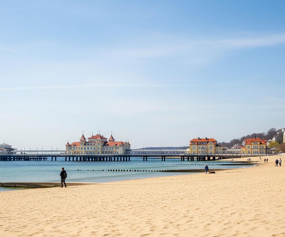 Piękny widok plaży z budynkami i molo w tle.
Dominującym elementem jest szeroka, piaszczysta plaża rozciągająca się od prawego dolnego rogu aż do lewej strony obrazu, wzdłuż turkusowej wody morskiej, gdzie widoczne są pojedyncze pale falochronów wystające z wody.

W tle, ponad wodą, znajduje się długie molo, na którym widać kilka budynków o żółtych fasadach i czerwonych dachach, z najbardziej okazałym budynkiem o licznych wieżyczkach pośrodku.

Na plaży widać kilka osób, w tym jedną w ciemnej kurtce z plecakiem, stojącą bliżej brzegu i patrzącą w stronę morza, oraz dwie inne idące w oddali w prawą stronę.

Na horyzoncie widoczne jest bezchmurne, jasnoniebieskie niebo, a po prawej stronie obrazu, za zabudowaniami, widać zbocze wzgórza porośnięte drzewami o nagich gałęziach.