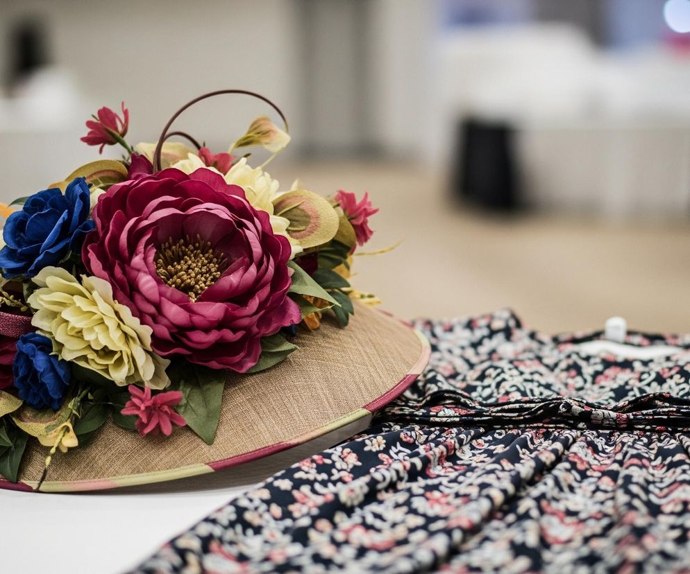 Na pierwszym planie leży ozdobne nakrycie głowy, przypominające toczek, w kolorze beżowym, z teksturowaną powierzchnią, obszyte różowo-zieloną lamówką. Jest ono bogato udekorowane sztucznymi kwiatami w odcieniach intensywnego różu, żółci i błękitu, oraz zielonymi liśćmi i brązowymi, zakręconymi elementami dekoracyjnymi. Obok toczka, po prawej stronie, leży złożona czarna tkanina z drobnym, kwiatowym wzorem w kolorach różowym, białym i beżowym. Tło jest rozmyte, widoczne są jasne powierzchnie oraz ciemne, niewyraźne kształty, sugerujące wnętrze pomieszczenia.