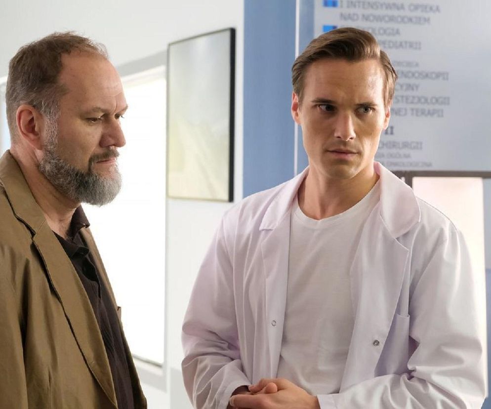 Mirosław Wójcik (Jakub Kornacki) i Olek Chodakowski (Maurycy Popiel) w scenie z serialu „M jak miłość”. Wójcik, mężczyzna z brodą w brązowej marynarce, patrzy na Olka w białym kitlu lekarskim. Obaj stoją w pomieszczeniu przypominającym poczekalnię w placówce medycznej. Więcej o ich losach na Super Seriale.