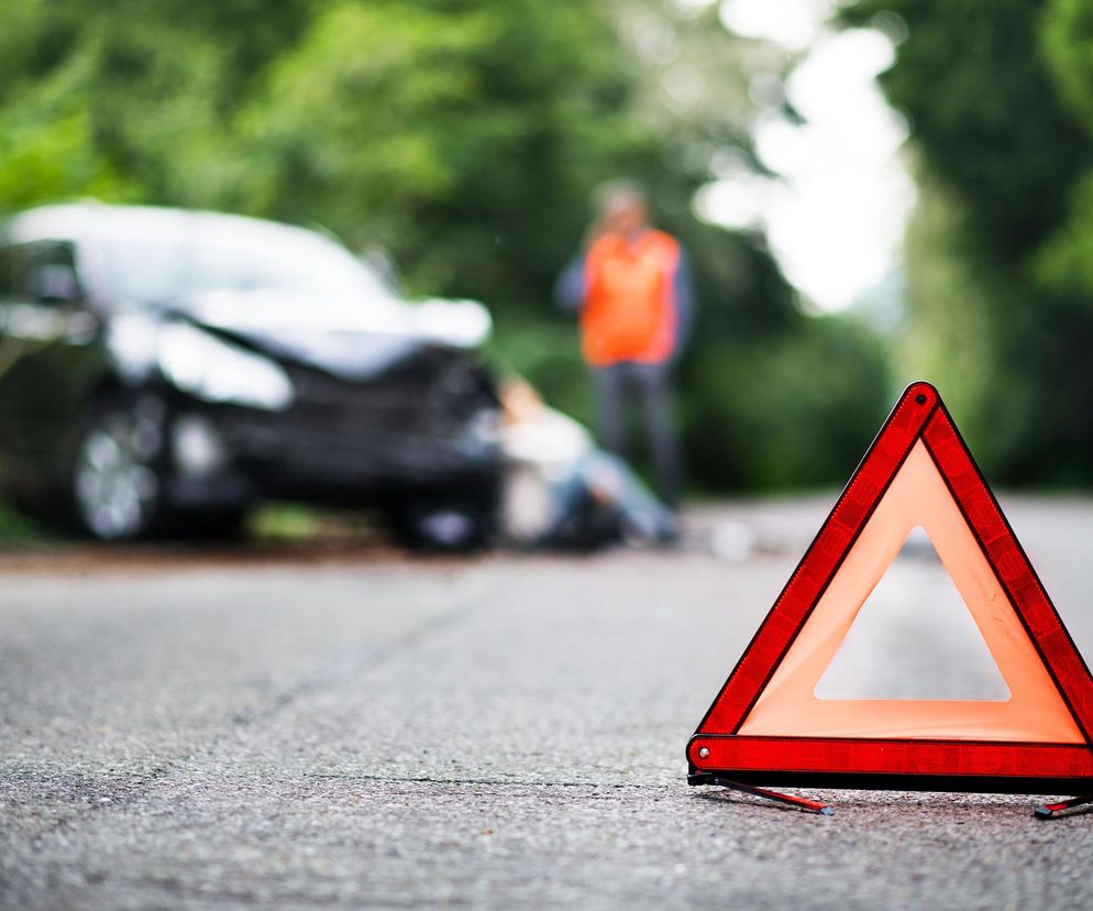 Trójkąt ostrzegawczy na pierwszym planie, w tle rozmazana scena wypadku drogowego z uszkodzonym ciemnym samochodem, leżącą postacią i osobą w kamizelce odblaskowej, obrazująca ryzyko prowadzenia pod wpływem. Więcej na ten temat przeczytasz na Poradnik Zdrowie.