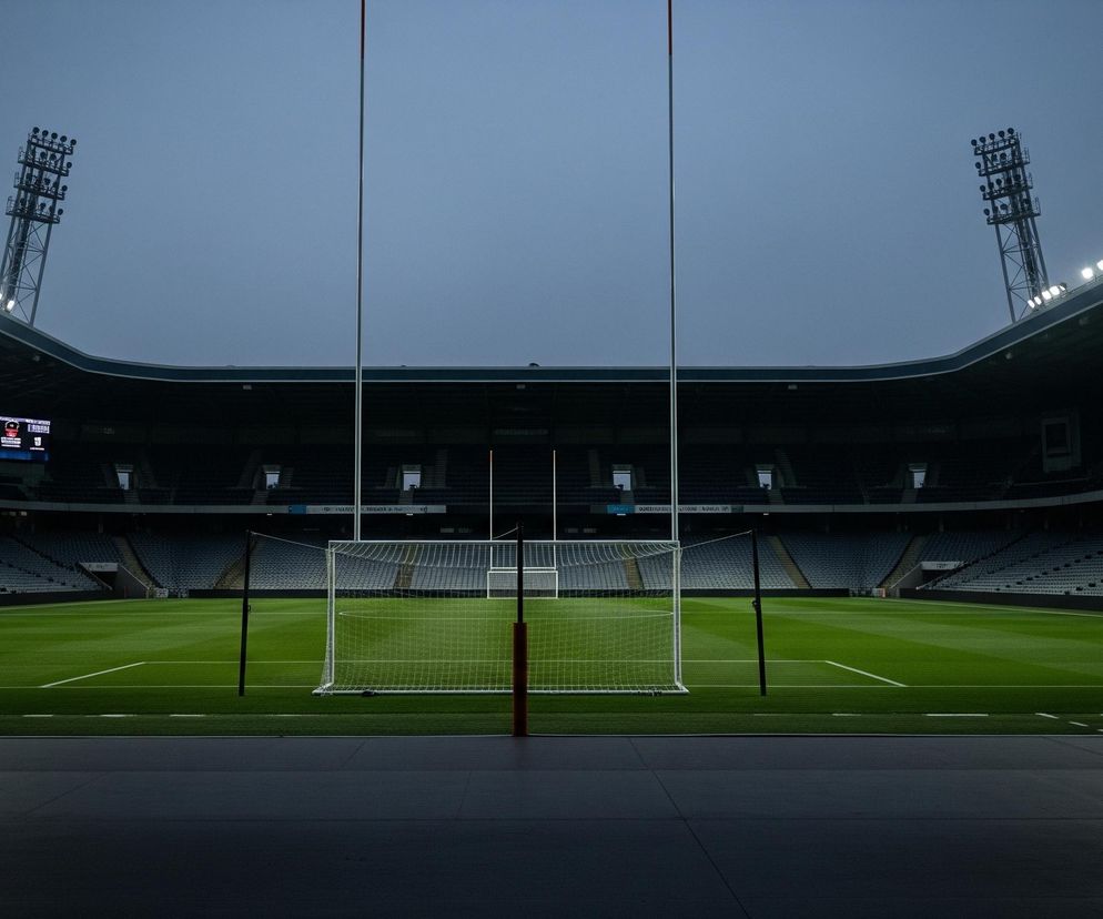 Pusty stadion sportowy rozciąga się w dali, wypełniony rzędami szarych siedzeń, które schodzą w kierunku zielonego boiska. Na pierwszym planie widać ciemny, betonowy obszar, a za nim murawę z białymi liniami, na której ustawiona jest biała bramka z siatką, flankowana przez dwie wysokie, białe słupy z czerwonymi akcentami. Niebo nad stadionem jest jasnoszare, a z obu stron stadionu widać zapalone jupitery, rozświetlające górne partie konstrukcji.