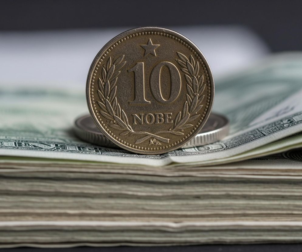 Na pierwszym planie, centralnie, stoi moneta w kolorze brązu lub starego złota, z wyraźnym cyframi 10 i napisem NOBE pod nimi, otoczonymi wieńcem z liści. U góry monety znajduje się pięcioramienna gwiazda, a jej krawędź jest ozdobiona drobnymi kropkami. Moneta ta stoi na zwiniętym stosie banknotów w odcieniach zieleni, bieli i szarości, które tworzą grubą warstwę falujących linii widocznych w dolnej części obrazu. W tle, częściowo widoczna jest inna, srebrzysta moneta, a całość tła jest rozmyta, ukazując jaśniejsze i ciemniejsze obszary, z dominującą ciemnoszarą barwą u góry.