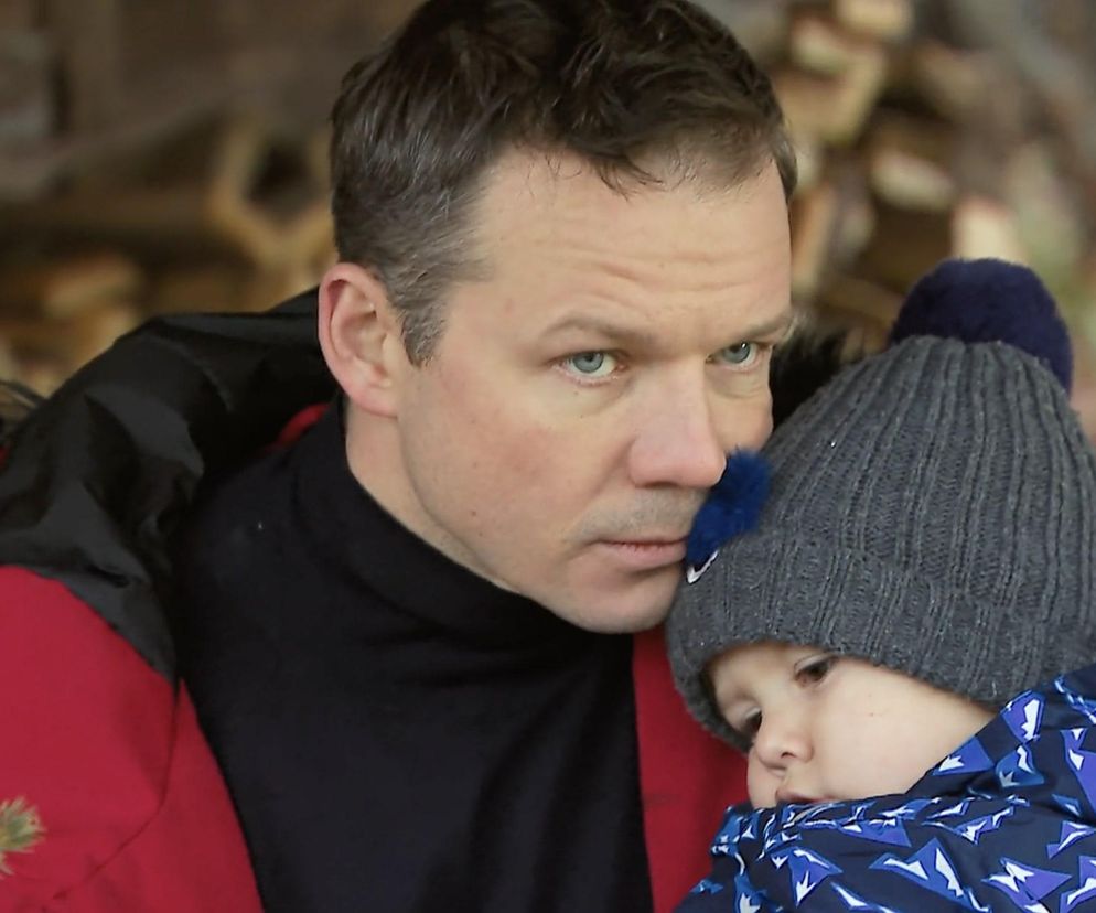 Bliskie ujęcie Bruna (Lesław Żurek) w czerwonej kurtce i czarnym golfie, patrzącego poważnie w dal. Tuli do siebie syna Tadzia (Józio Trojanowski) w niebieskiej kurtce i szarej czapce z pomponem. Zdjęcie ilustruje sceny z serialu Barwy szczęścia, o których można przeczytać na portalu Super Seriale.