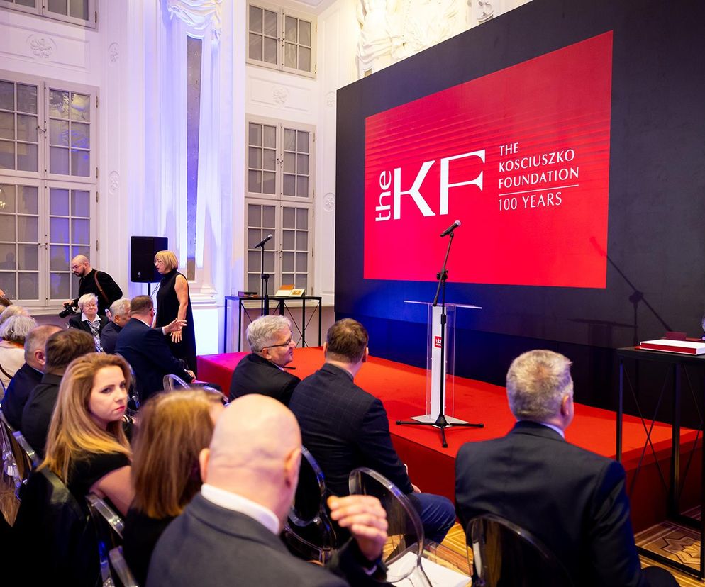 Uczestnicy gali z okazji 100-lecia Fundacji Kościuszkowskiej w Pałacu Rzeczypospolitej, siedzący w rzędach, obserwują scenę z czerwonym podestem. W tle duży ekran wyświetla logo Fundacji i napis 100 Years. Więcej o tym ważnym wydarzeniu można przeczytać na portalu Poradnik Zdrowie.