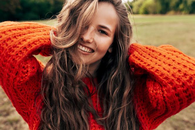 Uśmiechnięta młoda kobieta, która ubrana jest w pomarańczowy sweter z dzianiny. Pozuje na tle przyrody. 