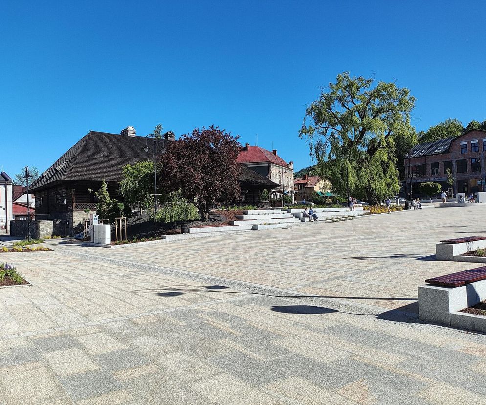 Sucha Beskidzka z perspektywy rynku, gdzie na pierwszym planie widoczne są ławeczki i nowocześnie wybrukowany plac. W tle góruje legendarna Karczma „Rzym” z charakterystycznym drewnianym dachem, obok której rozpościerają się zielone drzewa. Więcej o tym magicznym miejscu na naszym portalu.