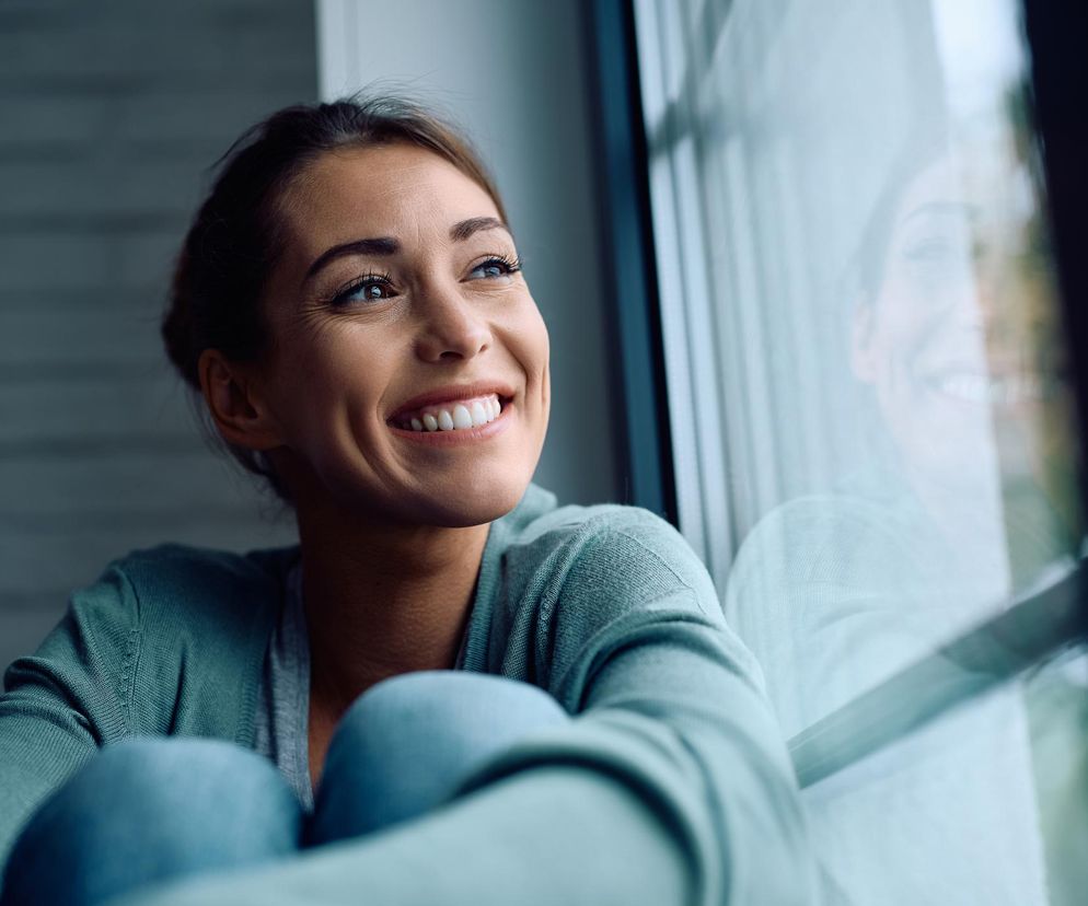 Uśmiechnięta kobieta z podwiniętymi kolanami, w zielonym swetrze, spoglądająca przez okno, z refleksami światła na szybie. Kobieta symbolizuje spokój i dobrą kondycję psychiczną, którą można osiągnąć dzięki zwiększeniu poziomu BDNF, o czym więcej przeczytasz na Poradnik Zdrowie.