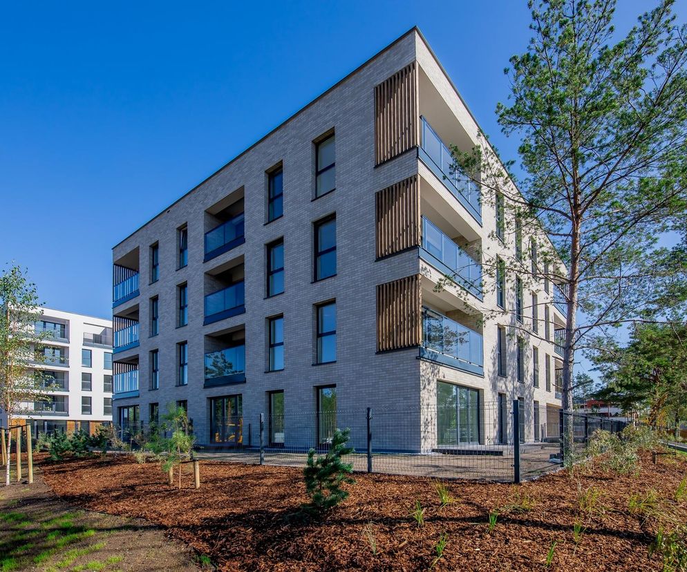 Nowoczesny budynek mieszkalny Naturia w Toruniu, z szarą elewacją i drewnianymi lamelami, wpisany w zieleń osiedla JAR. Jasne balkony z przezroczystymi balustradami, naturalne materiały, podkreślające harmonię z otoczeniem. Projekt Kuryłowicz & Associates zgłoszony do konkursu Życie w Architekturze, o czym przeczytasz na Architektura Murator Plus.