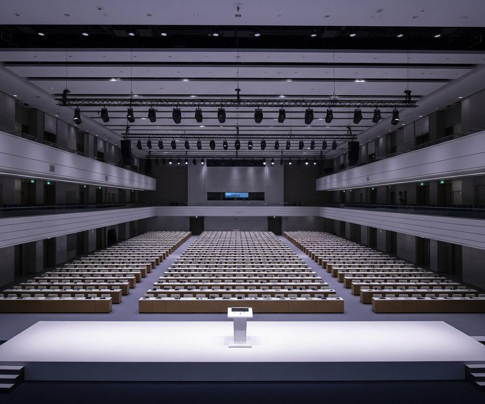 W przestronnej sali konferencyjnej, z centralnie umieszczonej perspektywy, dominuje duża biała scena z mównicą po prawej stronie. Przed sceną rozciąga się szereg długich, drewnianych stołów, ustawionych w równe rzędy z białymi krzesłami, zwróconymi w stronę sceny. Po obu stronach sali znajdują się balustrady balkonów, a na ścianach wzdłuż nich widoczne są zielone, podświetlone napisy. Nad całością rozciągają się konstrukcje kratownicowe z zamocowanymi reflektorami, a w oddali na końcu sali widać duży ekran projekcyjny.