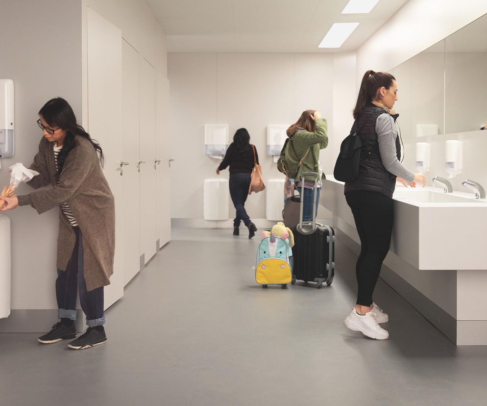 „Wielopokoleniowa scena w nowoczesnej, publicznej łazience. Matka pomaga dziecku wytrzeć ręce przy dozowniku do ręczników marki Tork, natomiast w tle inne kobiety korzystają  z umywalek i dozowników. Ilustracja kampanii „Inkluzywna Higiena” Tork, o której przeczytasz na Architektura Murator Plus.”