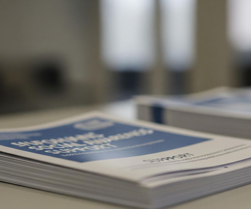 Na pierwszym planie leży stos broszur lub ulotek z biało-niebieskimi okładkami, częściowo otwarty. Na okładce widać biały tekst na niebieskim tle, zawierający słowa SOCIAL AWARENESS SUPPORT. Po lewej stronie stosu widoczne są niebieskie paski na grzbietach kartek. W tle, rozmyty, znajduje się drugi, podobny stos broszur.