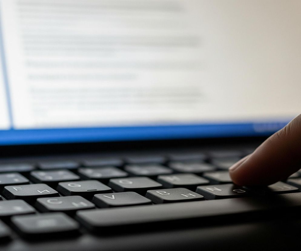 Na pierwszym planie widoczna jest ciemna klawiatura laptopa, a prawy palec wskazujący dotyka klawisza. Klawisze mają białe litery i cyfry, w tym D, F, B, H, N oraz symbole cyrylicy. Tło stanowią rozmyte treści wyświetlane na ekranie laptopa, gdzie dominują jasne obszary tekstowe i niebieski pasek na dole, z niewyraźnymi ikonami.