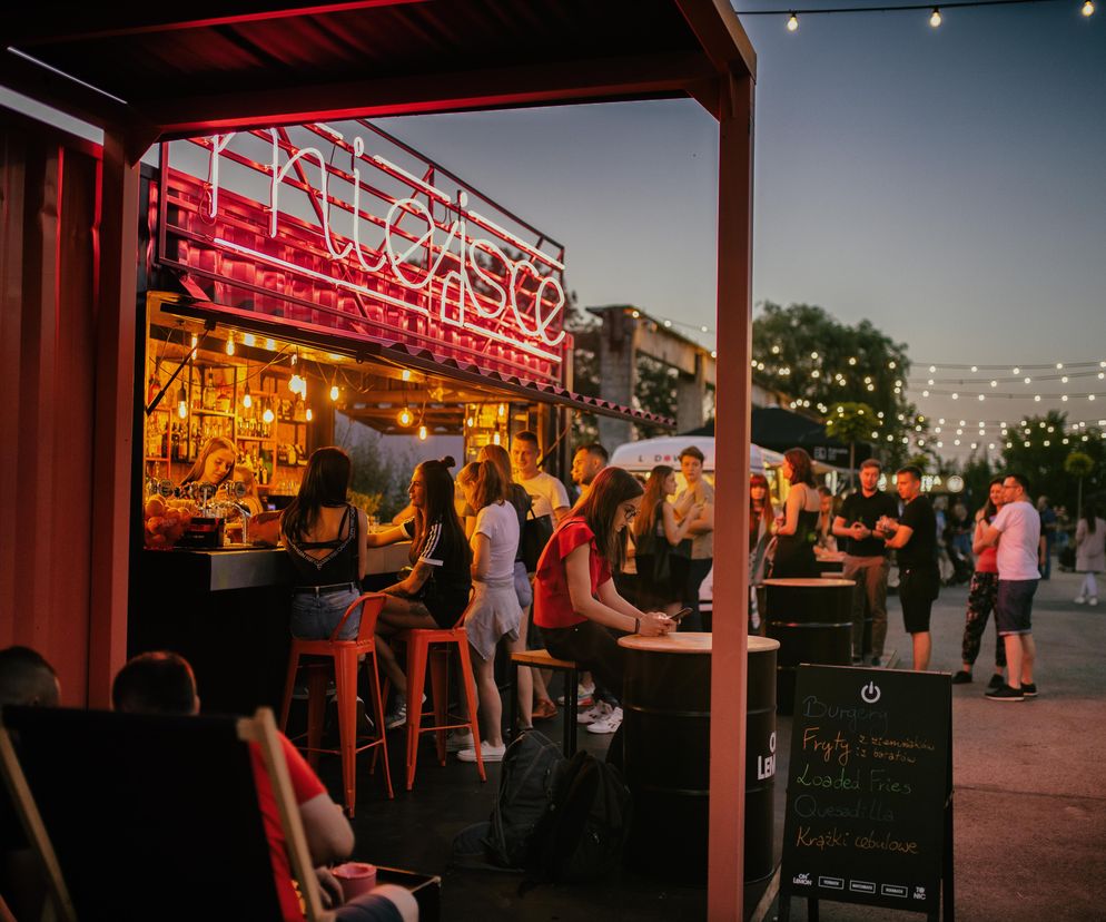 Wieczorne ujęcie lokalu „Miejsce” w Dąbrowie Górniczej, wykonanego z adaptowanych kontenerów morskich. Widoczne jest jaskrawe, neonowe logo lokalu, pomarańczowe elementy konstrukcji oraz girlandy świetlne. Tłum ludzi gromadzi się wokół baru i stolików, tworząc atmosferę spotkań. Więcej o adaptacjach przestrzeni publicznych przeczytasz na Architektura Murator Plus.