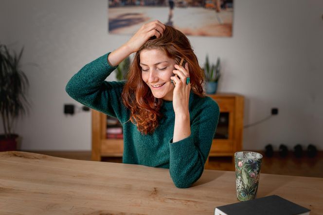 Uśmiechnięta kobieta siedzi przy stole w domu i rozmawia przez telefon