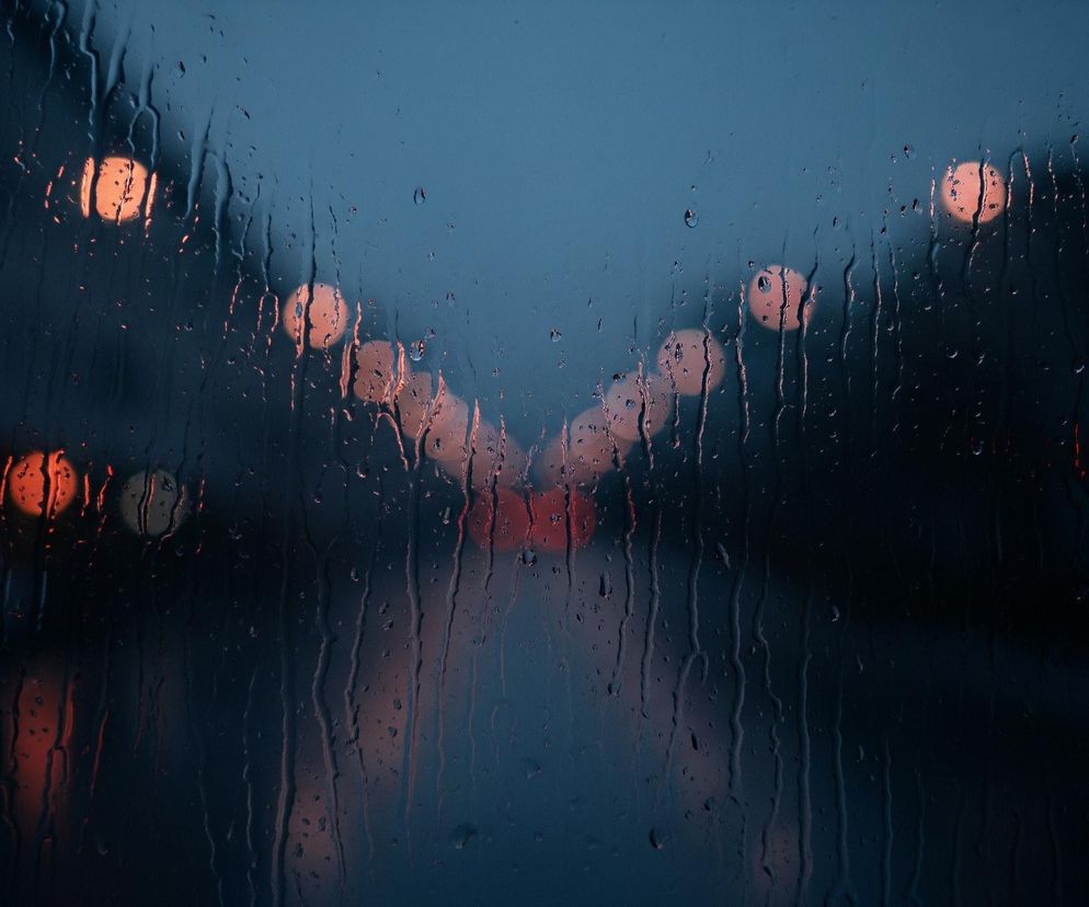 Szklana powierzchnia, prawdopodobnie szyba, jest gęsto pokryta deszczem, widocznym jako liczne krople i smugi wody spływające pionowo. W tle rozmazane są jasne, okrągłe punkty światła w odcieniach pomarańczowego i czerwonego, ułożone w perspektywiczny kształt litery V, zbiegający się ku dołowi i centrum obrazu. Całość utrzymana jest w ciemnej, chłodnej tonacji granatu i błękitu, z kontrastującymi ciepłymi światłami.