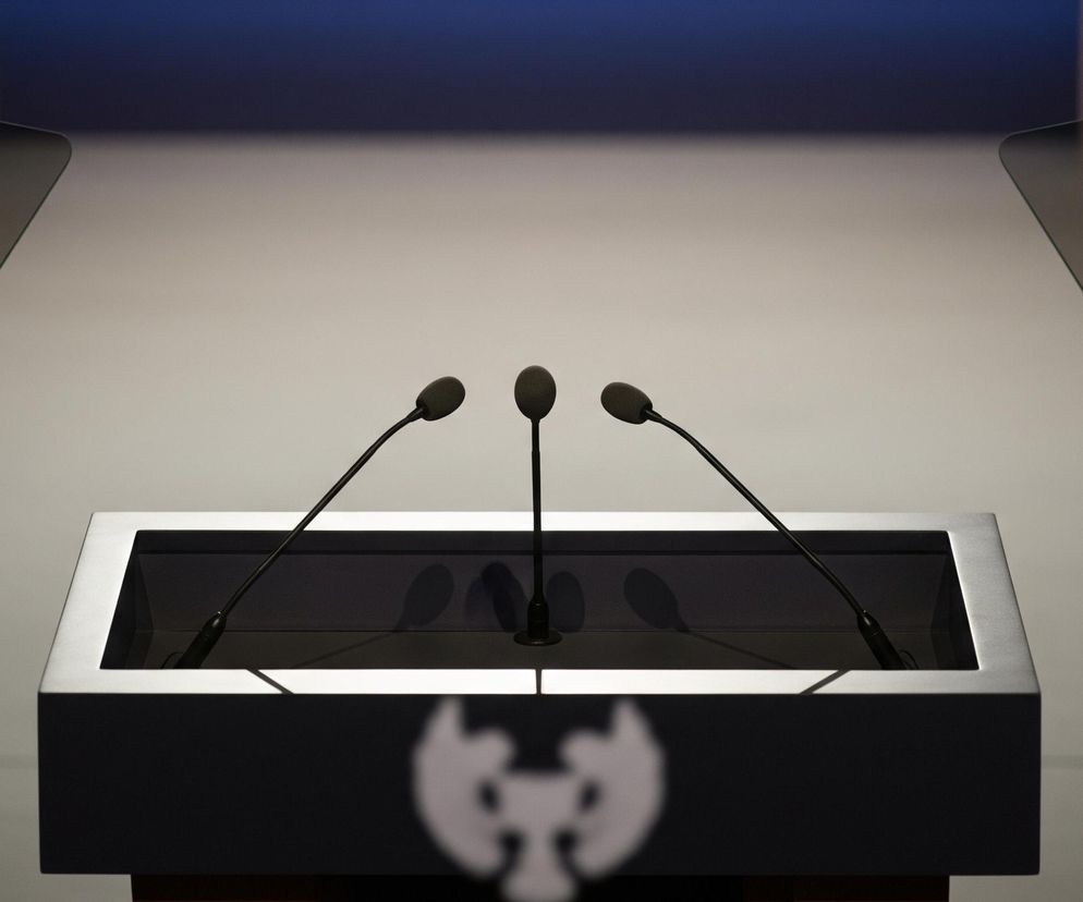 Centralnie umieszczona jest ciemna mównica z trzema czarnymi mikrofonami na cienkich, giętkich wysięgnikach, skierowanymi ku górze. Przednia część mównicy jest czarna, z niewyraźnym, białym logo w dolnej części. Po obu stronach mównicy stoją przezroczyste, ciemne ekrany teleprompterów na czarnych statywach. Tło jest rozmyte, w górnej części widać pas ciemnoniebieskiego koloru, przechodzący w jaśniejszy odcień szarości ku dołowi.
