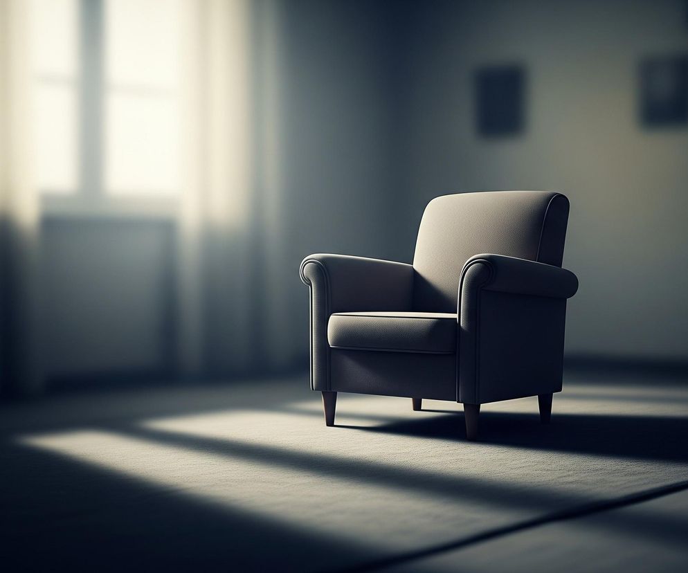 Beżowy fotel stoi na środku pokoju, oświetlony jasnymi promieniami słońca padającymi przez okno z lewej strony. Podłoga pokryta jest dywanem o szarym odcieniu, na którym widoczne są cienie tworzące wyraźne pasy. Ściany w pomieszczeniu są ciemniejsze, z niewyraźnymi, prostokątnymi kształtami na prawej ścianie.