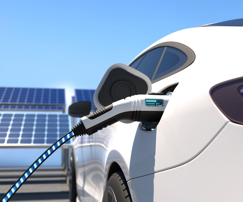 Biały samochód elektryczny ładowany z futurystycznej stacji, obok panele słoneczne. Na wtyczce widać wskaźnik ładowania 52% i dwie minuty do końca. To nowa era ładowania, o której przeczytasz na Super Biznes.