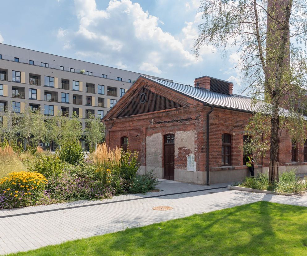 Fragment rewitalizowanego obszaru Fuzja w Łodzi, ukazujący zabytkowy ceglany budynek z nowoczesnym osiedlem mieszkaniowym w tle. Widoczna zielona przestrzeń miejska z bujną roślinnością i ścieżką dla pieszych, o której można przeczytać na Architektura Murator Plus.