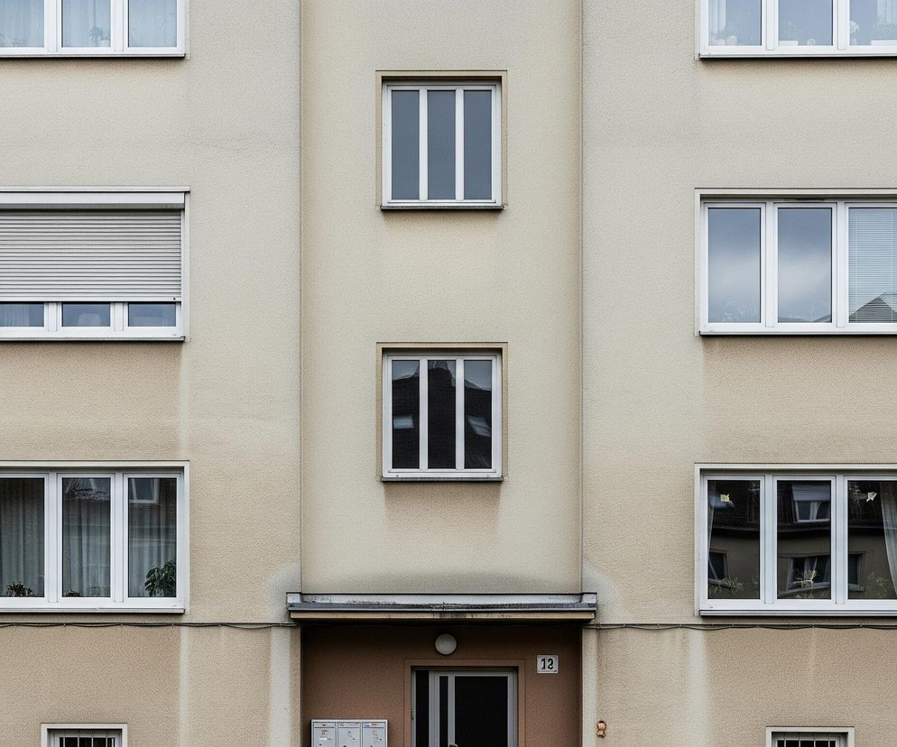 Centralną część obrazu zajmuje beżowa fasada budynku mieszkalnego, z licznymi oknami i elementami architektonicznymi. Po lewej i prawej stronie widoczne są fragmenty balkonów z metalowymi balustradami w kolorze brązowym. Na środku fasady, poniżej drugiego od góry okna, znajduje się wejście do budynku z brązowymi drzwiami i czarnym numerem 12 obok. Okna są prostokątne, w białych ramach, niektóre z nich posiadają żaluzje lub firanki, a w szybach odbija się otoczenie.