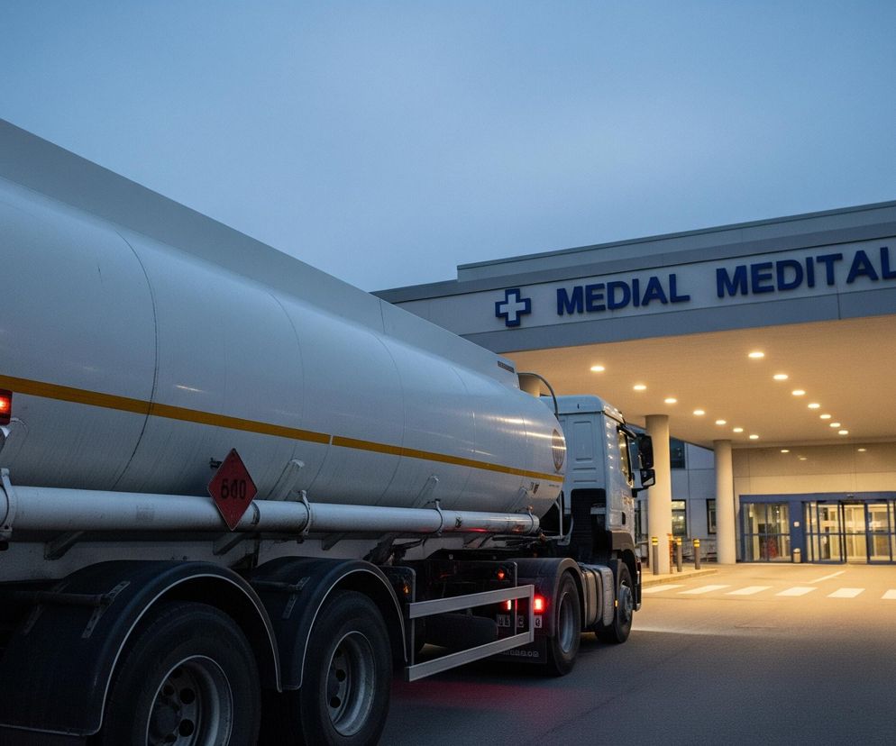 Na pierwszym planie, z lewej strony, dominuje biała cysterna ciężarówki, częściowo widoczna, skierowana w stronę centrum obrazu. Na jej boku widoczne są czerwone przerywane linie oraz romb z liczbą 640. Pojazd ma włączone tylne światła pozycyjne, emanujące czerwonym światłem. W tle znajduje się nowoczesny budynek z jasnym oświetleniem pod zadaszonym wejściem, nad którym widnieje napis MEDIAL MEDITAL z niebieskim krzyżem. Przed wejściem do budynku widoczne są białe pasy przejścia dla pieszych na ciemnym asfalcie. Całość oświetlona jest w tonacji zmierzchowej, z lekko niebieskim niebem w górnej części obrazu.