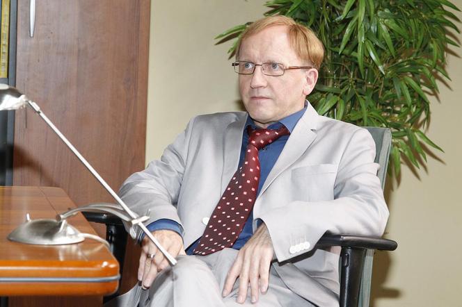 Aktor Artur Barciś w okularach, w jasnym garniturze i czerwonym krawacie z białymi kwadratami, siedzi na krześle przy biurku. Dowiaduj się więcej o powrocie Czerepacha z serialu „Ranczo” na naszym portalu.