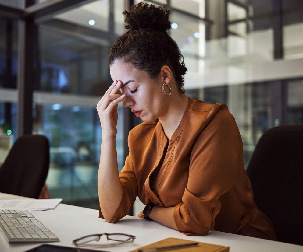 Młoda kobieta w biurowym stroju, z kędzierzawymi włosami upiętymi w kok, siedząca przy biurku w pracy, opiera czoło na dłoni, co sugeruje zmęczenie lub ból głowy. Scena ma miejsce wieczorem, a tło przedstawia biurowiec zza okna. Na Poradnik Zdrowie znajdziesz artykuły o wpływie stresu i odwodnienia na samopoczucie.