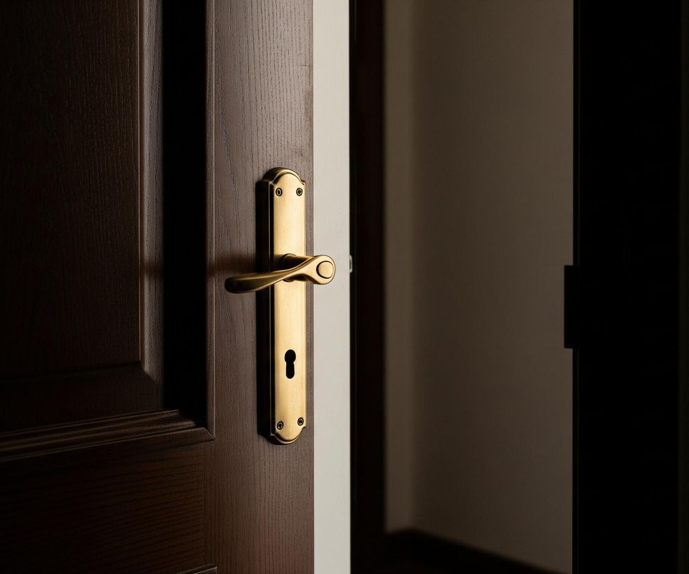 Na zdjęciu widoczna jest uchylona na prawo, ciemnobrązowa drewniana, fornirowana, gładka z widoczną fakturą drewna, skrzydło drzwiowe. Na drzwiach znajduje się złota, błyszcząca klamka z zamkiem na klucz, skierowana w prawo. Po prawej stronie drzwi, za nimi, widać ciemne wnętrze pomieszczenia. Kontrast między jasnym światłem padającym na klamkę a cieniem w pomieszczeniu tworzy dramatyczny efekt.