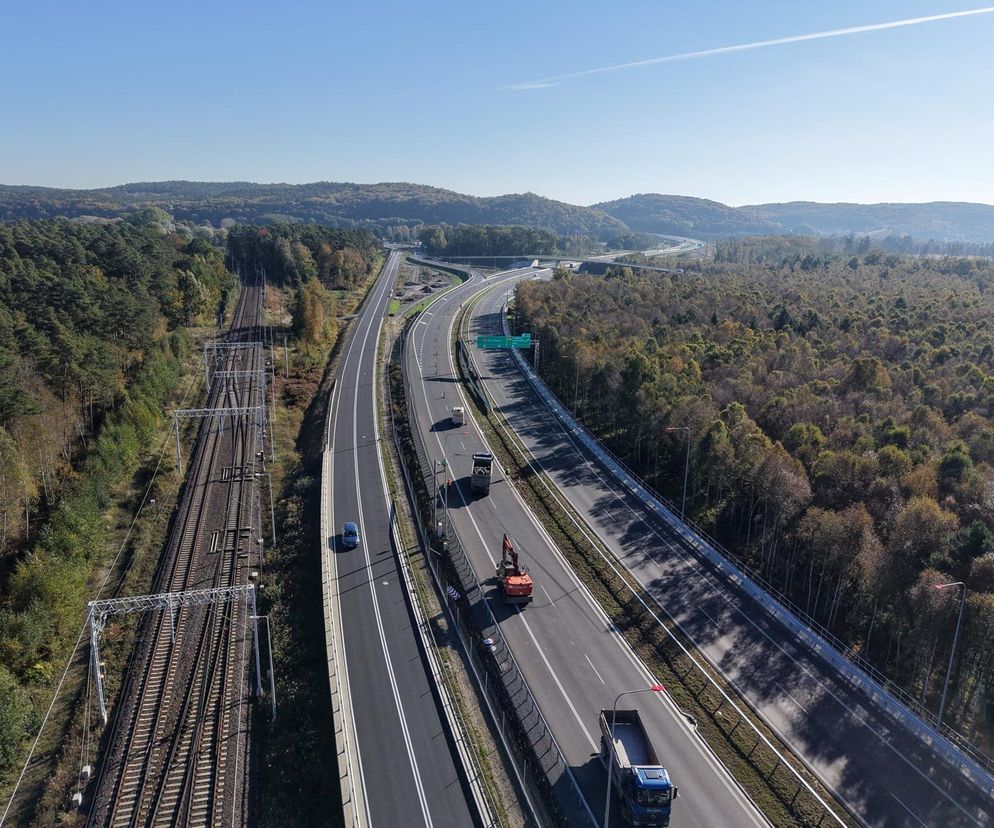 Widok z lotu ptaka na nowoczesną drogę ekspresową S3, biegnącą przez zalesione tereny, z widoczną infrastrukturą kolejową obok. Na dwupasmowej jezdni poruszają się samochody osobowe i ciężarowe. O otwarciu drogi przeczytasz na Super Biznes.