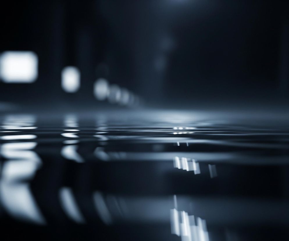 Powierzchnia wody, lekko pofałdowana, odbija rozmyte światła w oddali, tworząc długie, jasne smugi i punkty. Dominują ciemne odcienie szarości i błękitu, z jaśniejszymi, niemal białymi obszarami światła. W górnej części kadru widoczne są niewyraźne, podłużne kształty, sugerujące odległe źródła światła w ciemnym, przestrzennym wnętrzu. Całość utrzymana jest w niskim kluczu, z głęboką ostrością na falującej powierzchni.