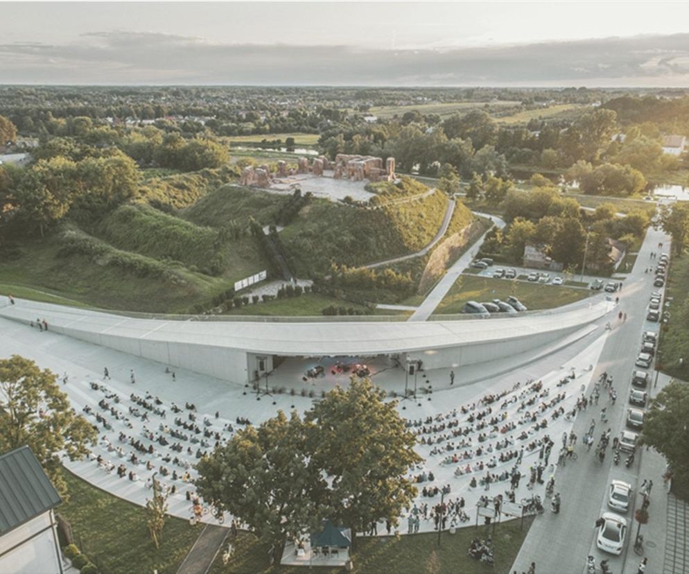 Widok z lotu ptaka na nowoczesny amfiteatr z widownią i sceną otwartą na ruiny zamku na wzgórzu oraz tereny zielone wzdłuż rzeki Bzury w Sochaczewie, co jest idealnym przykładem zagospodarowania przestrzeni publicznej na portalu Architektura Murator Plus.