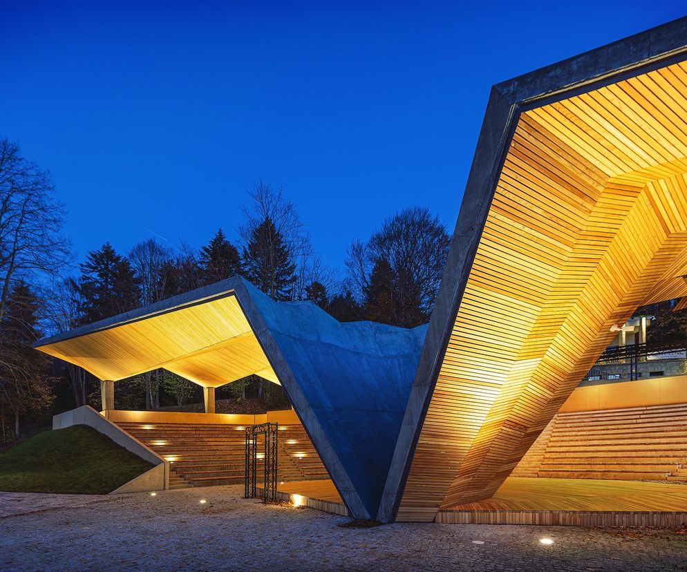 Nowoczesny amfiteatr w Krynicy-Zdroju autorstwa Saneccy sp. z o.o. Architektura x Krajobraz, oświetlony ciepłym światłem po zmierzchu, z drewnianym zadaszeniem i betonowymi elementami, wpisuje się w otoczenie Parku Zdrojowego, o czym możesz przeczytać na Architektura Murator Plus.
