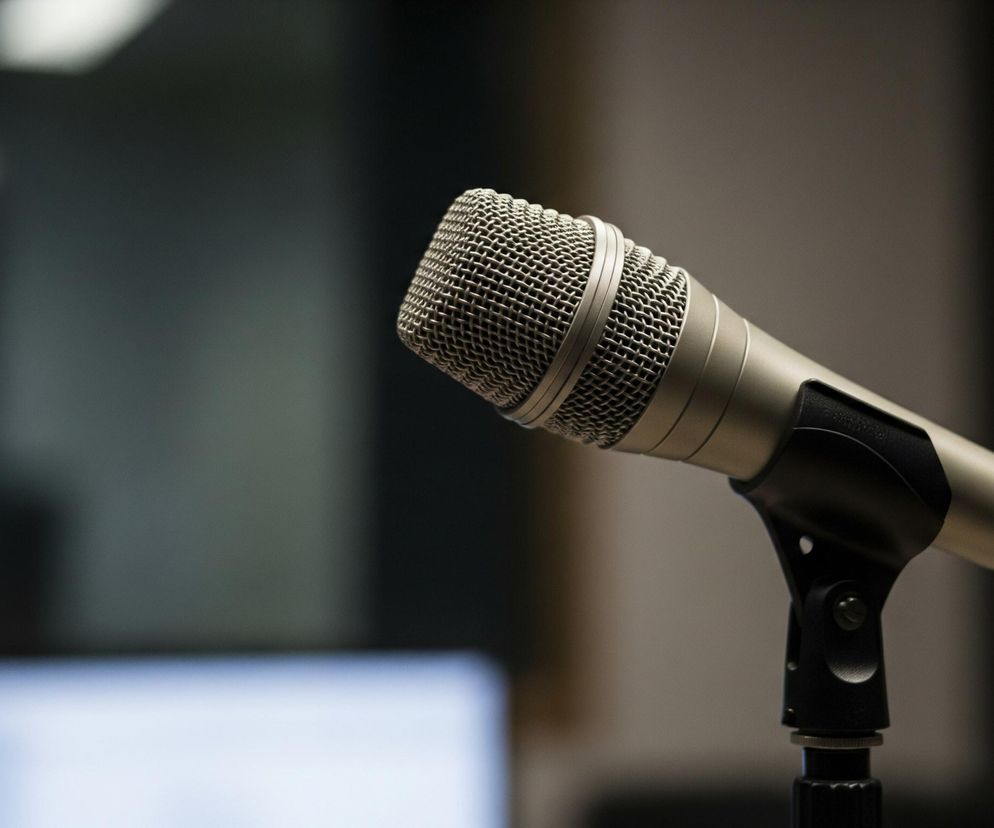 Srebrny mikrofon z gęstą metalową siatką na główce stoi na czarnym statywie, skierowany lekko w prawo i w dół. Korpus mikrofonu jest błyszczący, metaliczno-szary, a jego dolna część łączy się z czarnym przewodem. Tło jest rozmyte, dominują ciemne odcienie szarości i brązu, z kilkoma jasnymi punktami i fragmentem ekranu laptopa po lewej stronie.