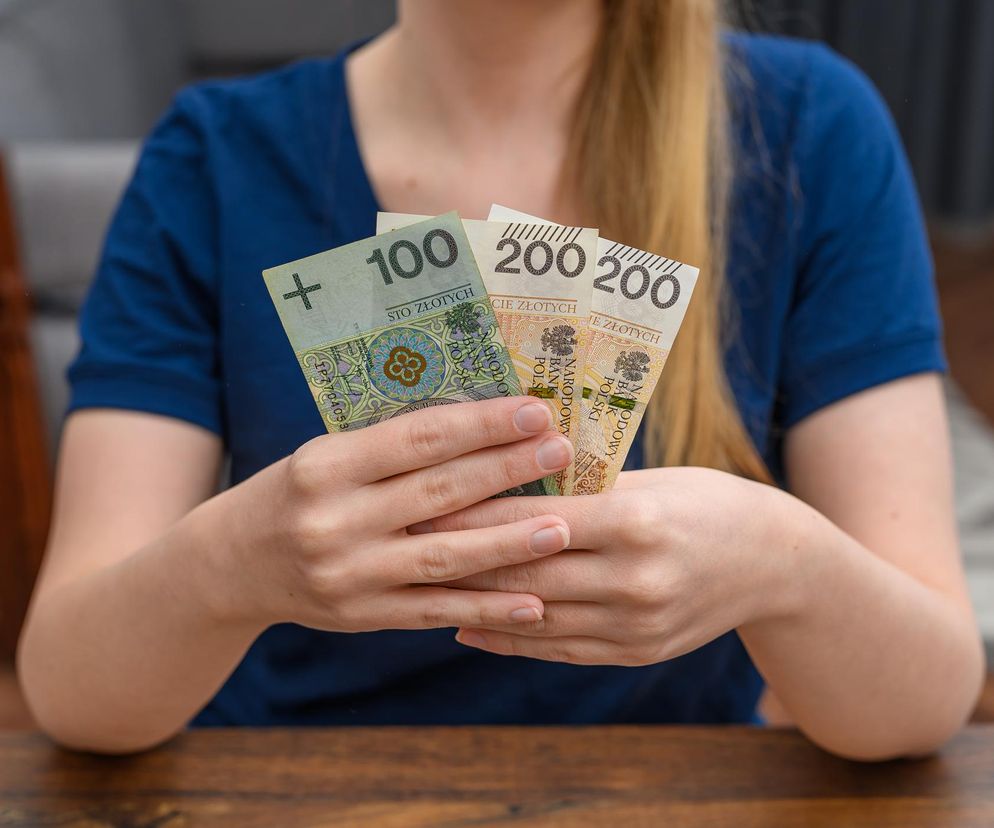 Kobieta trzyma w dłoniach polskie banknoty: jeden o nominale 100 złotych i dwa po 200 złotych, symbolizujące pełnopłatny urlop macierzyński oraz świadczenia finansowe dla rodziców. Więcej o nowym prawie pracy i zasiłkach macierzyńskich przeczytasz na Poradnik Zdrowie.