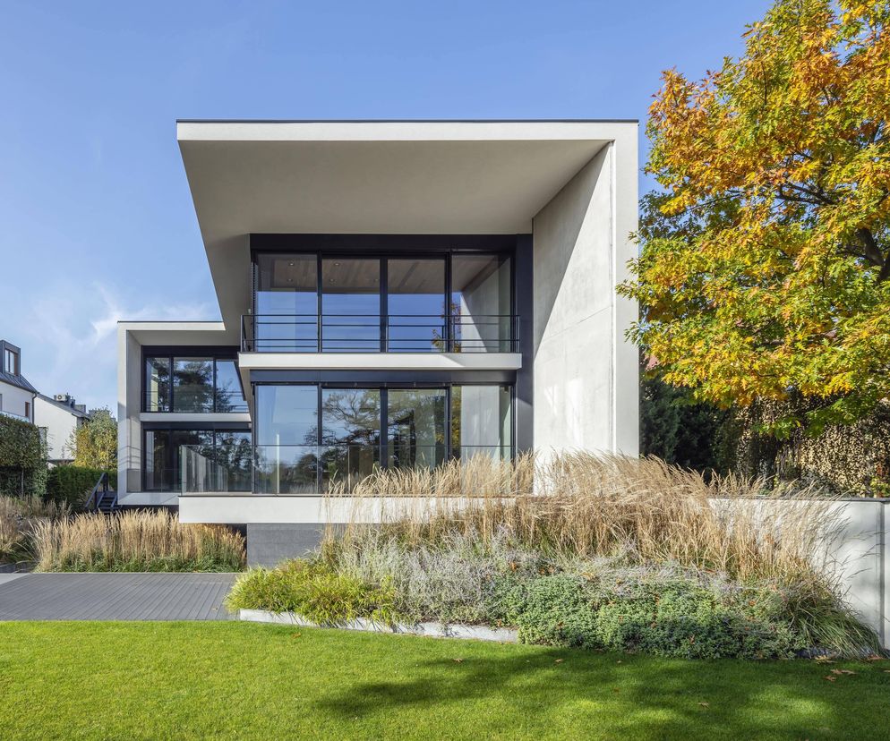 Nowoczesny, betonowy budynek mieszkalny Dom w Parku Cietrzewia pracowni Kruszewski Architekci z dużymi przeszkleniami i bujną roślinnością ozdobną, otoczony zielonym trawnikiem i jesiennym drzewem. Więcej o zgłoszeniu do konkursu Życie w Architekturze można przeczytać w Architektura Murator Plus.