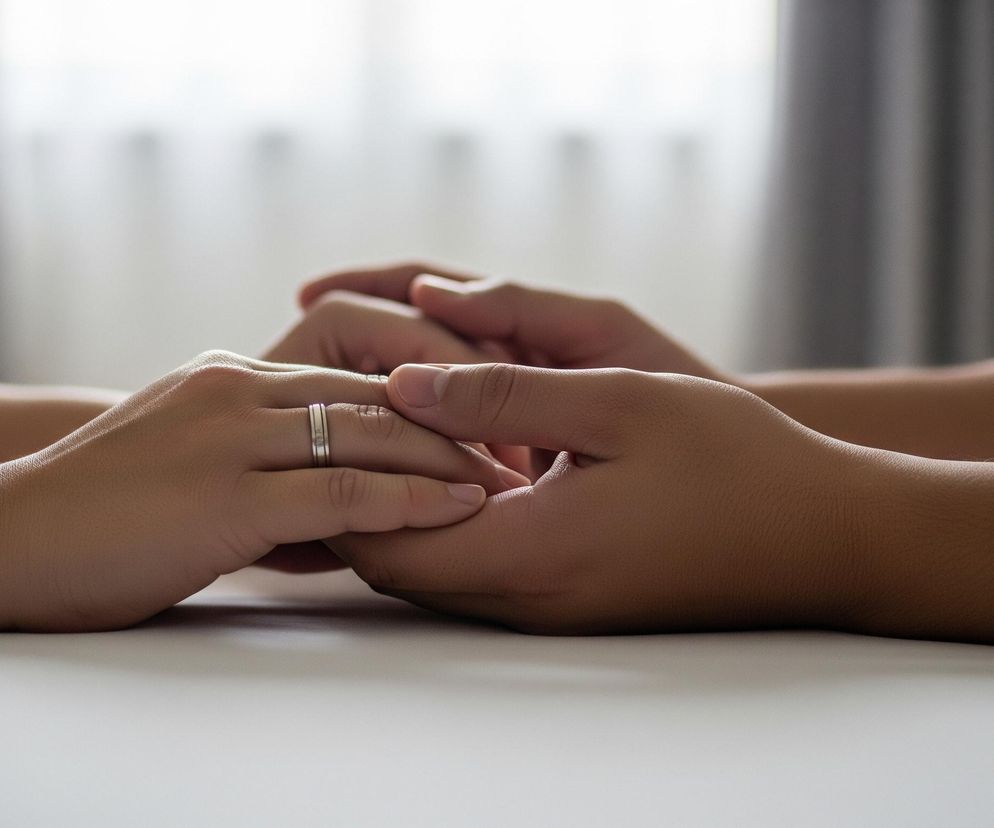 Dwie dłonie o różnym odcieniu skóry, ułożone na białej powierzchni, obejmują się nawzajem; lewa dłoń, jaśniejsza, ma na palcu obrączkę. Na obu dłoniach widać drobne zmarszczki i strukturę skóry. Tło jest rozmyte, z niewyraźnymi ciemnymi pionowymi kształtami po bokach i jaśniejszym obszarem pośrodku.